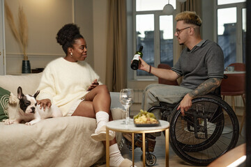 Side view of young man using wheelchair suggesting smiling African American girlfriend drinking bottle of red wine together celebrating happy relationship in cozy living room