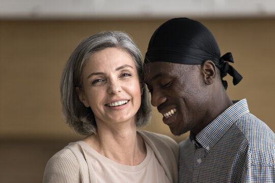 Beauty of love. Close up headshot portrait cheerful mature Caucasian woman looking at camera with happy smile standing close to beloved Black man partner who touch his forehead to her with affection