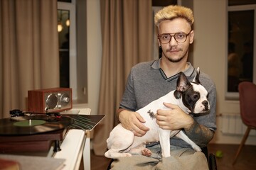 Portrait in warm tones of calm adult man using wheelchair holding adorable pied French bulldog on...