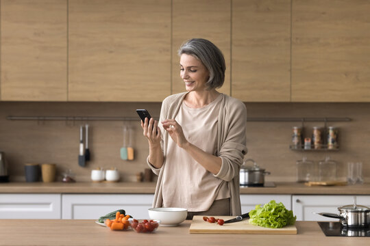Smart eating. Confident mid aged woman wife using mobile phone app to find healthy salad recipe search meal idea online in culinary blog when cooking natural vegetable food at home kitchen. Copy space