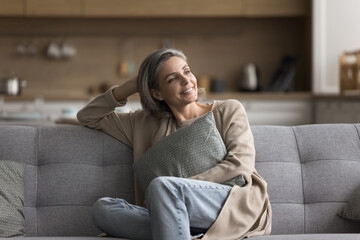 Happy at home. Carefree beautiful mature lady sit on sofa at comfortable pose hug pillow look away enjoy mindfulness moment relax alone on weekend planning pleasant life changes imagine house of dream