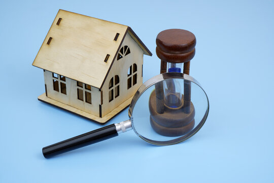 Wooden house model and hourglass under magnifying glass on blue background.
