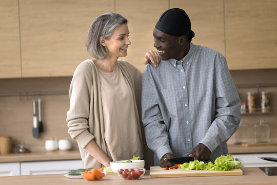 Home cooking with love. Happy bonding middle aged multiethnic family couple prepare healthy vegan salad for breakfast hug smile chat cutting fresh vegetables enjoy teamwork at stylish domestic kitchen