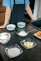 Unrecognizable young pastry chef is arranging bowls of flour, sugar, butter, and eggs on a sleek, dark countertop in a contemporary kitchen, ready to begin baking