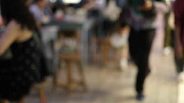 A blurry image of a restaurant with people sitting at tables and a man walking by. Scene is casual and relaxed, as people are enjoying their time at the restaurant