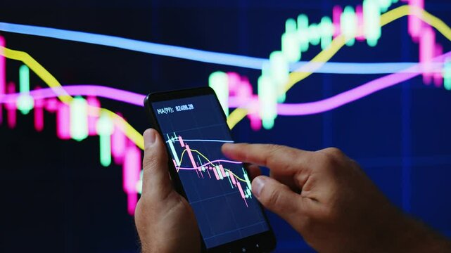 Trader or investor monitoring or looking stock market or crypto market graphics or charts on phone and PC. Man&rsquo;s hand holding phone with graphics in front of PS screen.