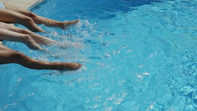 Girls are sitting at the edge of the pool with their legs dangling in the water and shaking the water  and making ripples and small waves. Summer background. 