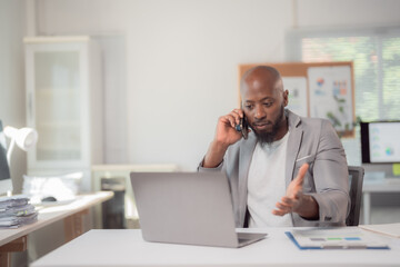 Obraz premium Focused african american businessman wearing a gray jacket and white t-shirt is sitting at his desk. Talking on his phone and working on his laptop in a bright office. While gesturing with his hand