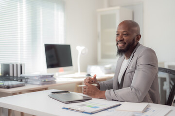 Fototapeta premium Smiling businessman sitting at a white desk, working on a laptop while holding a pen. Surrounded by a smartphone, papers, and files in a bright, modern office space