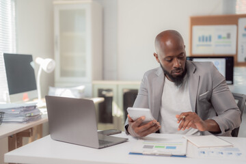 Focused African American businessman sitting at a modern office desk, using a calculator to check expenses, with a laptop and paperwork nearby, illustrating productivity