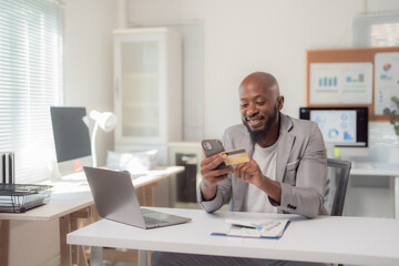 Businessman is sitting at his desk in a modern office, smiling while entering his credit card information on his phone to make an online purchase