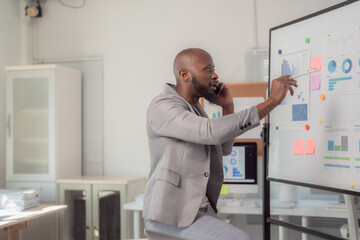 Fototapeta premium African American businessman in a gray suit sitting and pointing at a whiteboard with graphs while discussing marketing data and strategy on the phone in a modern office