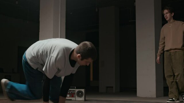 Handheld shot of inspired Caucasian gen Z b-boy performing headspin breakdance moves in front of peer, practicing indoors to boombox music