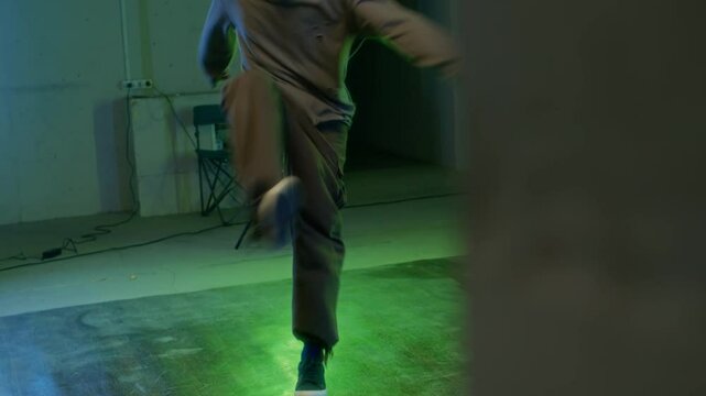 Young creative Caucasian b-boy with beard in urbanwear performing top rock and down rock moves of breakdance while practicing indoors under green and blue vibrant light