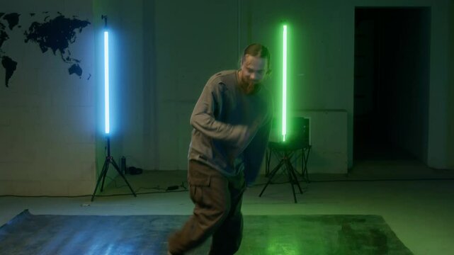 Zoom-in shot of young bearded Caucasian breaker in streetwear practicing top rock, improvising creative moves while breakdancing indoors under green and blue lamps
