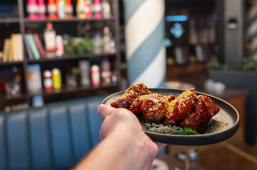 A hand holds a plate of delicious chicken wings coated in a rich glaze, garnished with herbs. The background features a stylish restaurant interior with colorful condiments.