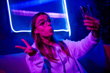 Photo of cute charming blonde girl take selfie photo v-sign sit sofa indoors nightclub neon blue purple light late night blogger
