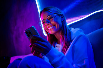 Photo of gamer lovely blonde young lady hold telephone sit sofa blogging neon light neon purple...