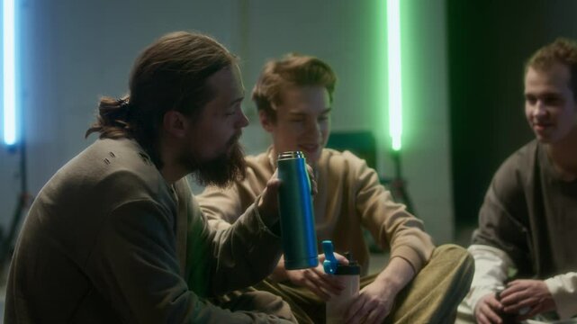 Slowmo of Caucasian male zoomers in urbanwear chatting with young Caucasian bearded man drinking water from sport bottle, chilling out together on floor during break in room with neon lighting