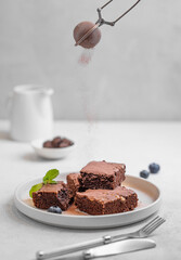 Cocoa powder pouring on chocolate brownie with nuts cut into pieces on plate on light background