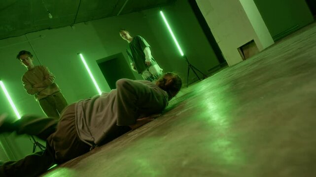 Ground level shot of young skilled Caucasian man in streetwear breaking in front of male zoomers indoors under green led lighting
