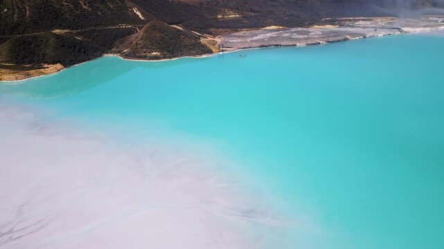 A turquoise tailings pond near a cement factory, surrounded by rugged hills. The vivid water contrasts with the barren land, creating a striking yet industrial scene of environmental impact