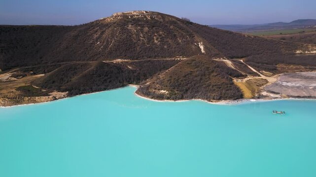 A turquoise tailings pond near a cement factory, surrounded by rugged hills. The vivid water contrasts with the barren land, creating a striking yet industrial scene of environmental impact