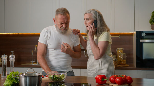 Ill sick Caucasian grandpa old senior male feeling bad at kitchen unhealthy unwell elderly man pain in chest heart attack heartburn heartache mature woman wife help husband call emergency with phone