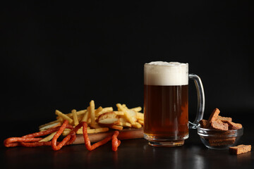 Glass with light beer and snacks on a dark background. Beer, French fries and hunting sausages. Glass on a wooden board and snacks. Light beer and bread croutons. Traditional glass beer mug