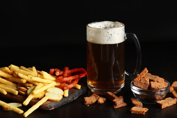 Glass with light beer and snacks on a dark background. Beer, French fries and hunting sausages. Glass on a wooden board and snacks. Light beer and bread croutons. Traditional glass beer mug