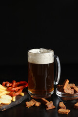 Glass with light beer and snacks on a dark background. Beer, French fries and hunting sausages. Glass on a wooden board and snacks. Light beer and bread croutons. Traditional glass beer mug