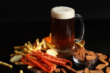 Glass with light beer and snacks on a dark background. Beer, French fries and hunting sausages. Glass on a wooden board and snacks. Light beer and bread croutons. Traditional glass beer mug