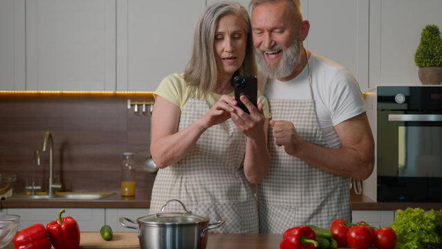 Happy senior old couple love bonding family adults woman man browsing smartphone online order delivery fresh food search recipe breakfast on mobile phone retired wife husband talk cook at home kitchen