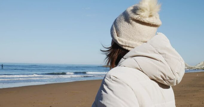 Mujer con gorro y chaqueta de abrigo llega a la playa al en los primeros d&iacute;as de primavera en un d&iacute;a soleado, y se quita el gorro y la chaqueta, frente al mar. Mientras su pero corre en el agua.