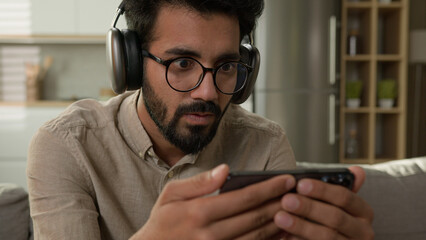 Shocked amazed Arabian man Indian guy watch video stream online sport match social media addict focused male in headphones looking at mobile phone screen watching movie on smartphone at home kitchen