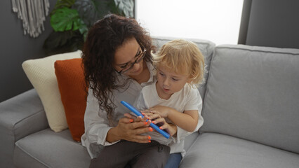 Woman and young boy sitting together on a sofa indoors, enjoying time using a tablet in a cozy living room, highlighting family bonding and modern technology use.