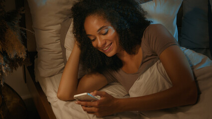African American happy woman addicted female in bed nighttime mobile phone scrolling networks social media smartphone gadget addict smiling ethnic girl browsing internet dating apps in night bedroom