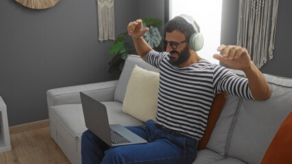Young man with a beard enjoying music through headphones while sitting on a couch with a laptop in a cozy living room.