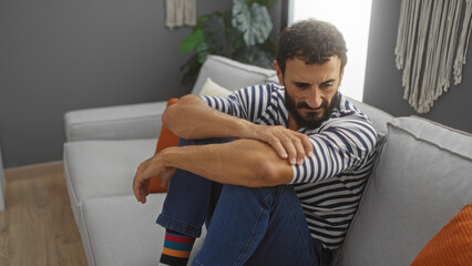 Young hispanic man sitting on a couch in a modern living room, wearing striped clothes and colorful...