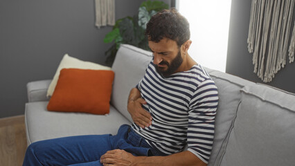 Young man with a beard feeling discomfort holding his stomach while sitting on a couch in a modern living room with striped shirt and indoor plants in the background