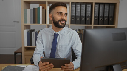 Handsome young hispanic man with beard holding tablet and sitting at desk in modern office workplace with bookshelves and documents in background