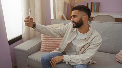 Fototapeta premium Young man taking selfie in living room with neutral expression on a cozy sofa, demonstrating casual indoor home environment with a modern aesthetic.