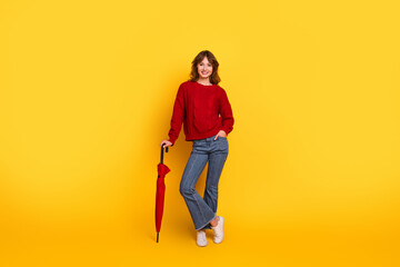 Fototapeta premium Smiling woman posing in red sweater and jeans holding an umbrella against a vibrant yellow background