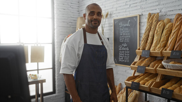 Young man in a bakery stands in front of a shelf of bread, wearing a blue apron, with a chalkboard menu in the background and a window letting in natural light.
