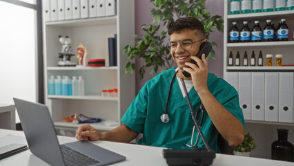 Young man wearing teal scrubs smiling and talking on the phone in a modern hospital room while...