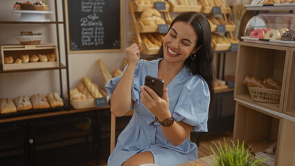Young woman celebrates good news while using her phone in a beautiful bakery filled with fresh bread and pastries