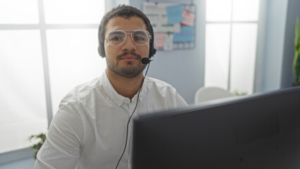 Young hispanic man wearing headset and glasses working in a bright office environment with a computer monitor in the foreground.