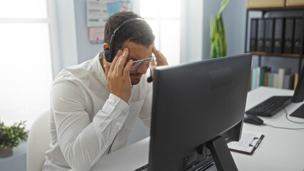 Young hispanic man wearing headset and glasses looks stressed, seated at a desk in a modern office...