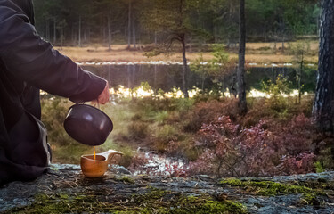 Pouring coffee from a kettle into a wooden cup near a lake in the forests
