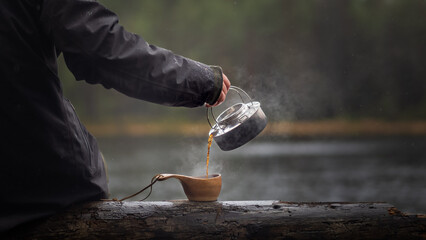 Pouring coffee from a kettle into a wooden cup near a lake in the forests on a rainy day